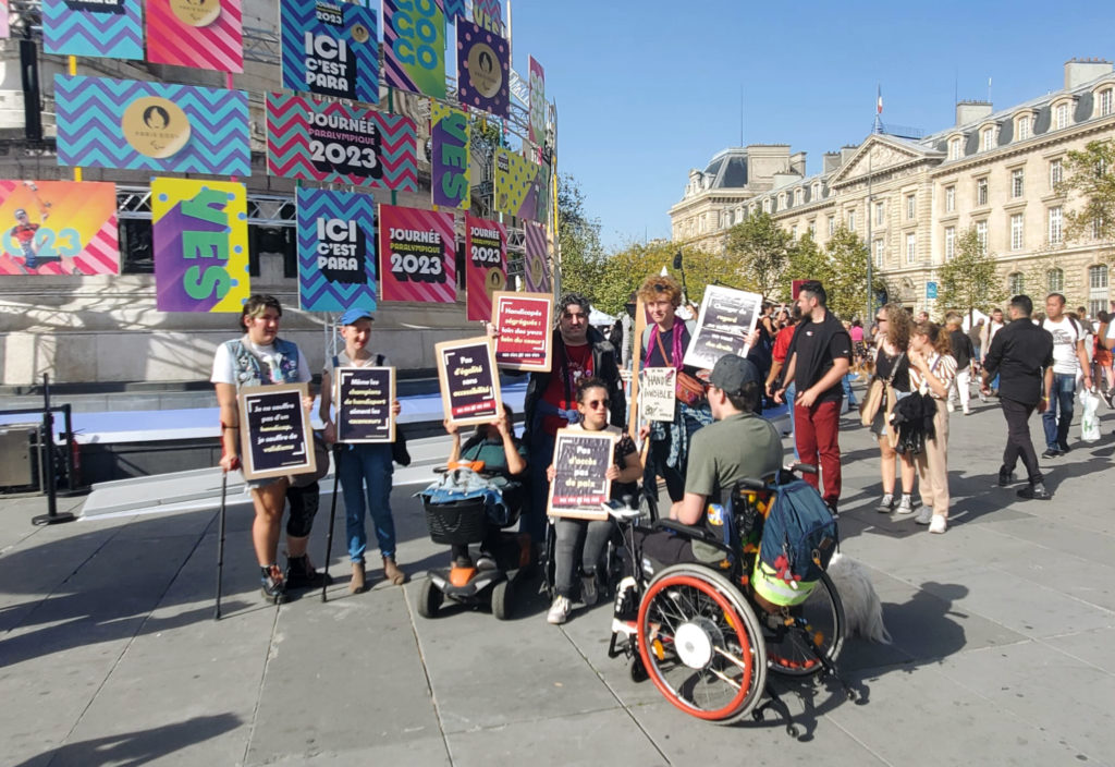 Sur la place de la République à Paris, devant des affiches colorées pour promouvoir la Journée Paralympique, un groupe de sept personnes handicapées tiennent des pancartes avec des slogans anti validistes, tels que "pas d'égalité sans accessibilité". L'homme en fauteuil roulant au premier plan tourne le dos à l'objectif et semble discuter avec le reste du groupe. 