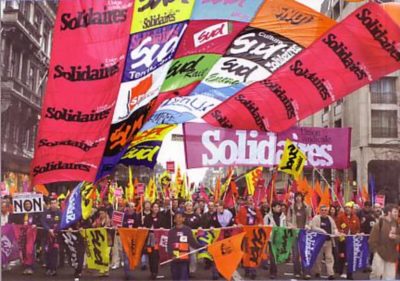 Photo d'une manifestation. De nombreuses banderoles multicolores volent dans les airs. On peut voir écrit Solidaire, ainsi que Sud et d'autres noms ne sont pas lisibles. Au premier plan, une rangée de personnes dans une manifestation tient une banderole multicolore horizontalement. 