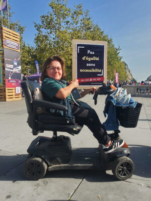 En plan rapproché, une femme handicapée en fauteuil roulant. Elle a les cheveux châtains coupés court et porte des lunettes. Elle tient dans ses mains une pancarte où il est écrit "Pas d'égalité sans accessibilité". Derrière, on aperçoit de grands arbres sur fond de ciel bleu, des drapeaux colorés et une foule de gens en contrebas. 