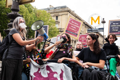 Photo d'un groupe des personnes, majoritairement des femmes, portant des pancarte "qui sème l'innacessibilité récolte la colère", "même les champions aiment les assensceurs". Elles tiennent une banderole qu'on ne voit pas en entier. 