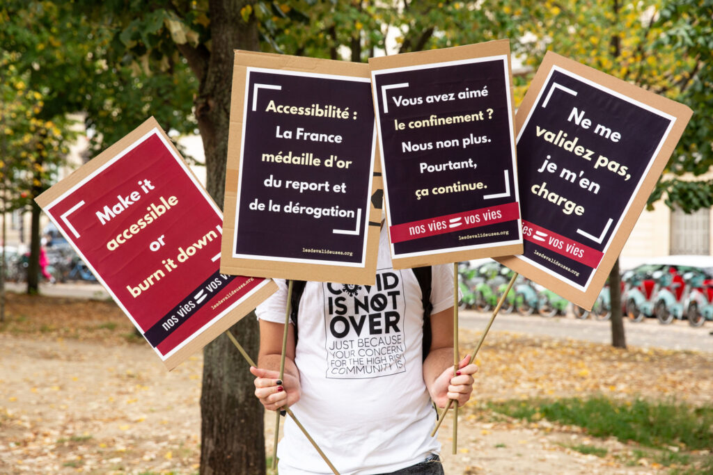 Une personne en tee-shirt blanc avec marqué "covid is not over" porte quatre pancartes roses et violettes. Sur la pancarte tout à gauche il est écrit "Make it accessible or burn it down". Sur la seconde pancarte : "Accessibilité, la France médaille d'or du report et de la dérogation". Sur la troisième pancarte : "Vous avez aimé le confinement ? Nous non plus. Pourtant, ça continue". Sur la quatrième pancarte : "Ne me validez pas, je m'en charge". 
