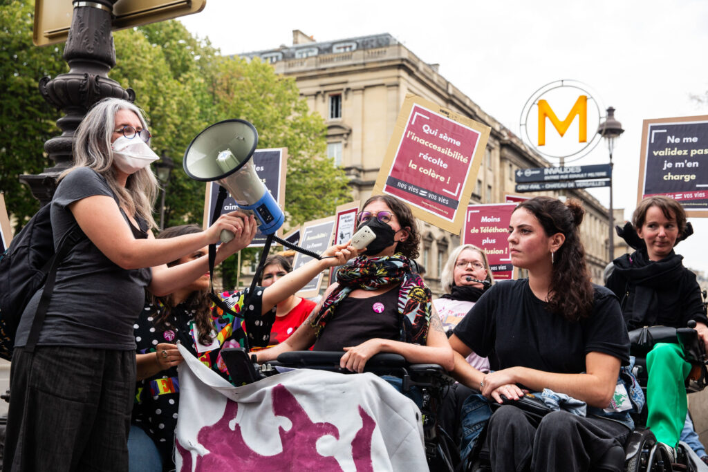 Plan rapproché sur des femmes handicapées, positionnées devant la station de métro des Invalides. Tout à gauche, une femme masquée tient un mégaphone pour la femme à côté d'elle, elle aussi masquée, assise sur un fauteuil roulant. Une troisième jeune femme au fauteuil complète cette première rangée ; elle se tient droite et attentive. Derrière, on voit d'autres femmes en fauteuil roulant, et des pancartes avec des slogans anti validistes tels que "Ne me validez pas, je m'en charge". 