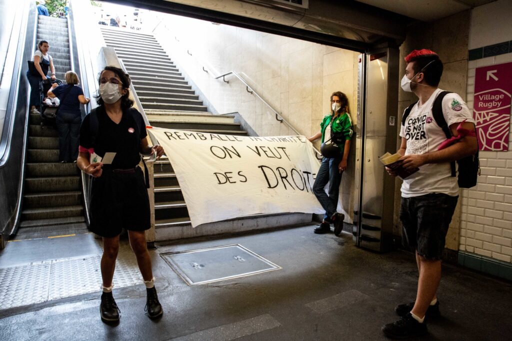 Trois personnes se tiennent en bas des escaliers d'une station de métro. L'ambiance de la photo est plutôt sombre. La femme à gauche est masquée et a des stickers dans les mains. L'homme tout à droite, également masqué, porte un tee-shirt blanc où il est marqué "covid is not over" et distribue des tracts. Juste derrière, au pied des marches, une jeune femme avec une veste verte tient une banderole qui bloque le passage vers les escaliers. Dessus il est écrit en grandes lettres noires : remballe ta charité, on veut des droits. 